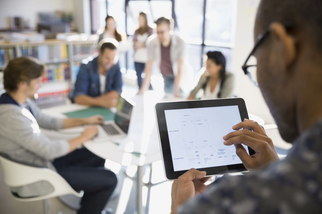 groepje mensen aan het werk in een bibliotheek rond een tafel met als achtergrond boekenkasten. Op de voorgrond staat iemand die werkt op een tablet