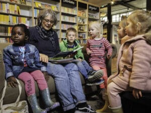 Een dame zit met verschillende kinderen op een bank in de bibliotheek en leest de kinderen voor.