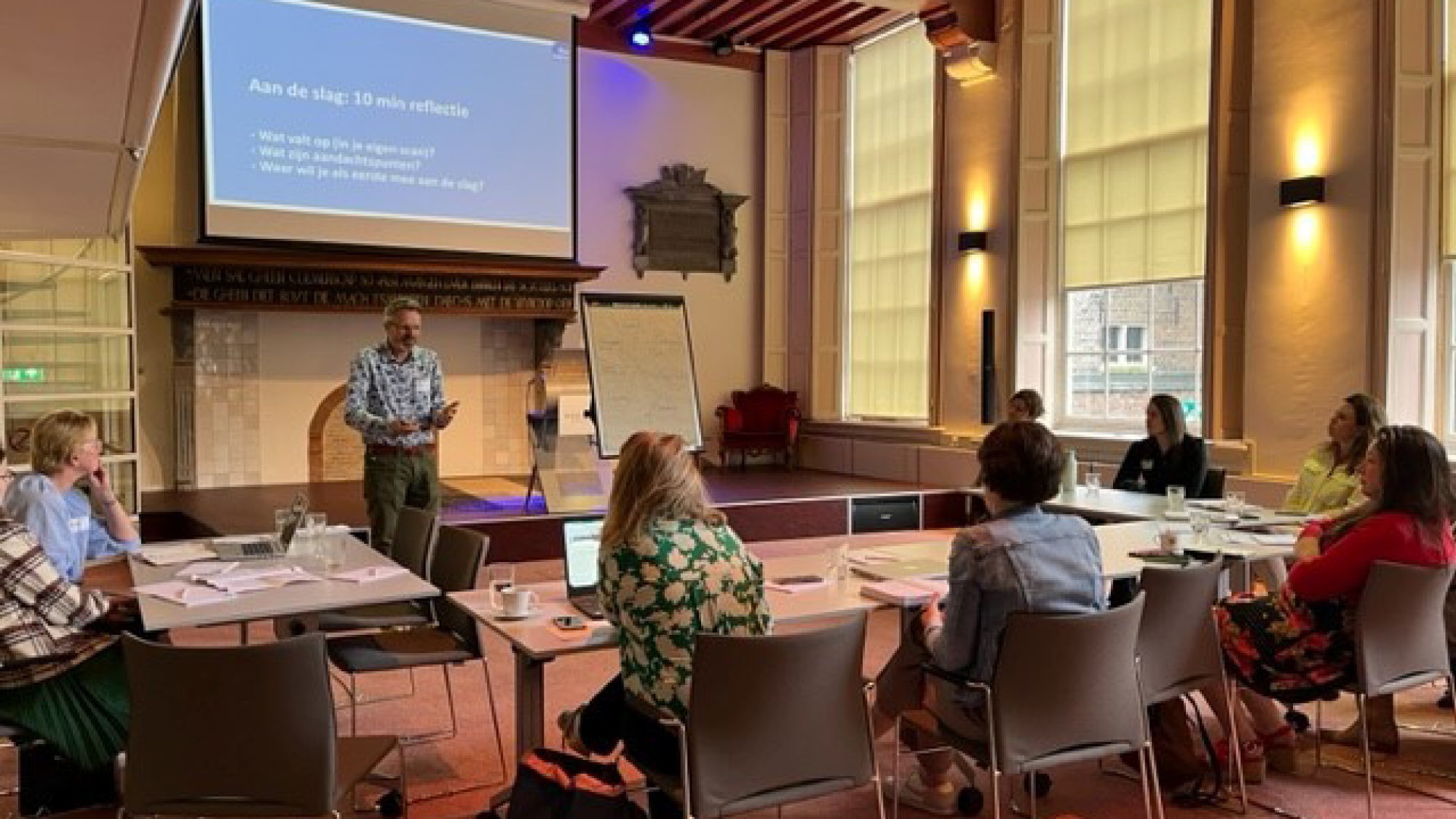 Een presentator spreekt een groep toe voor een groot scherm